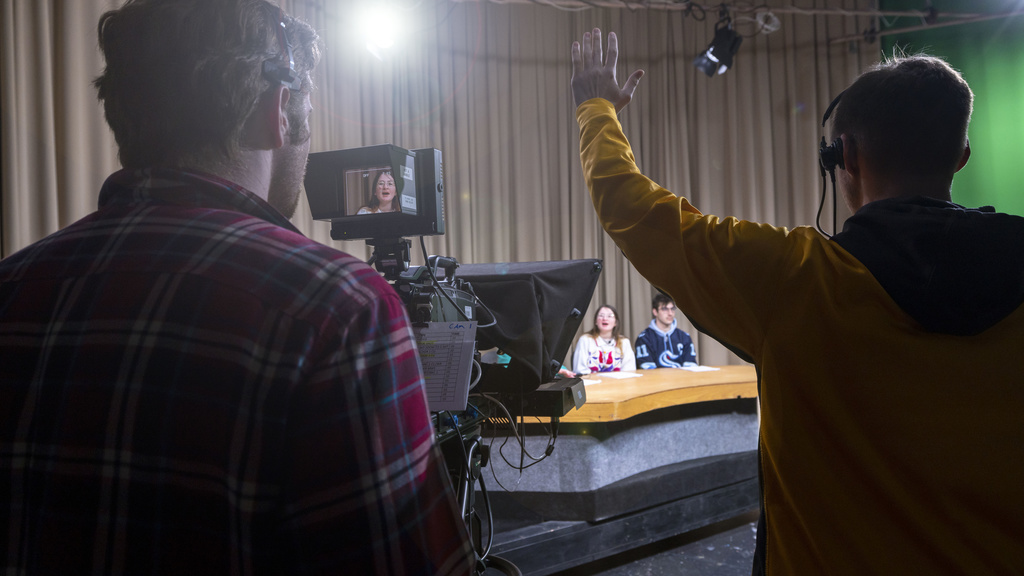 Students sitting at an anchor desk and standing behind the camera in a video production studio
